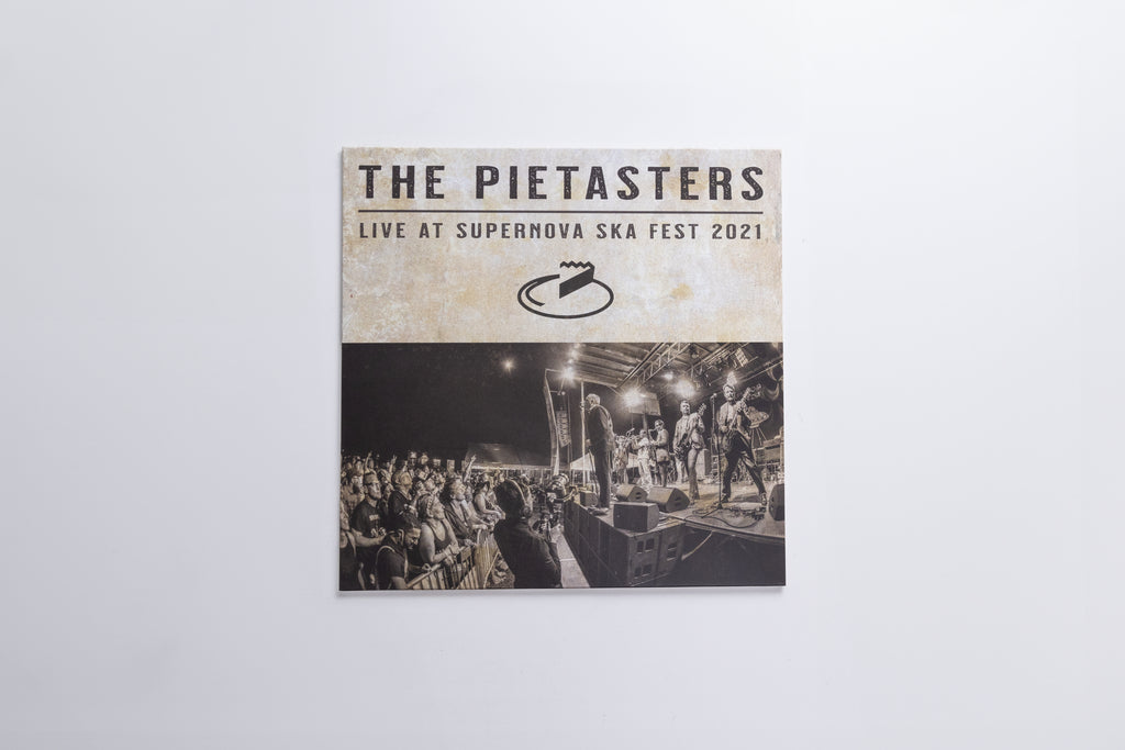 A square printed cover or poster for 'The Pietasters Live at Supernova Ska Fest 2021' hangs against a plain light gray wall. The top section is cream with black text and logo, while the bottom section shows a black-and-white concert scene with a stage and crowd.