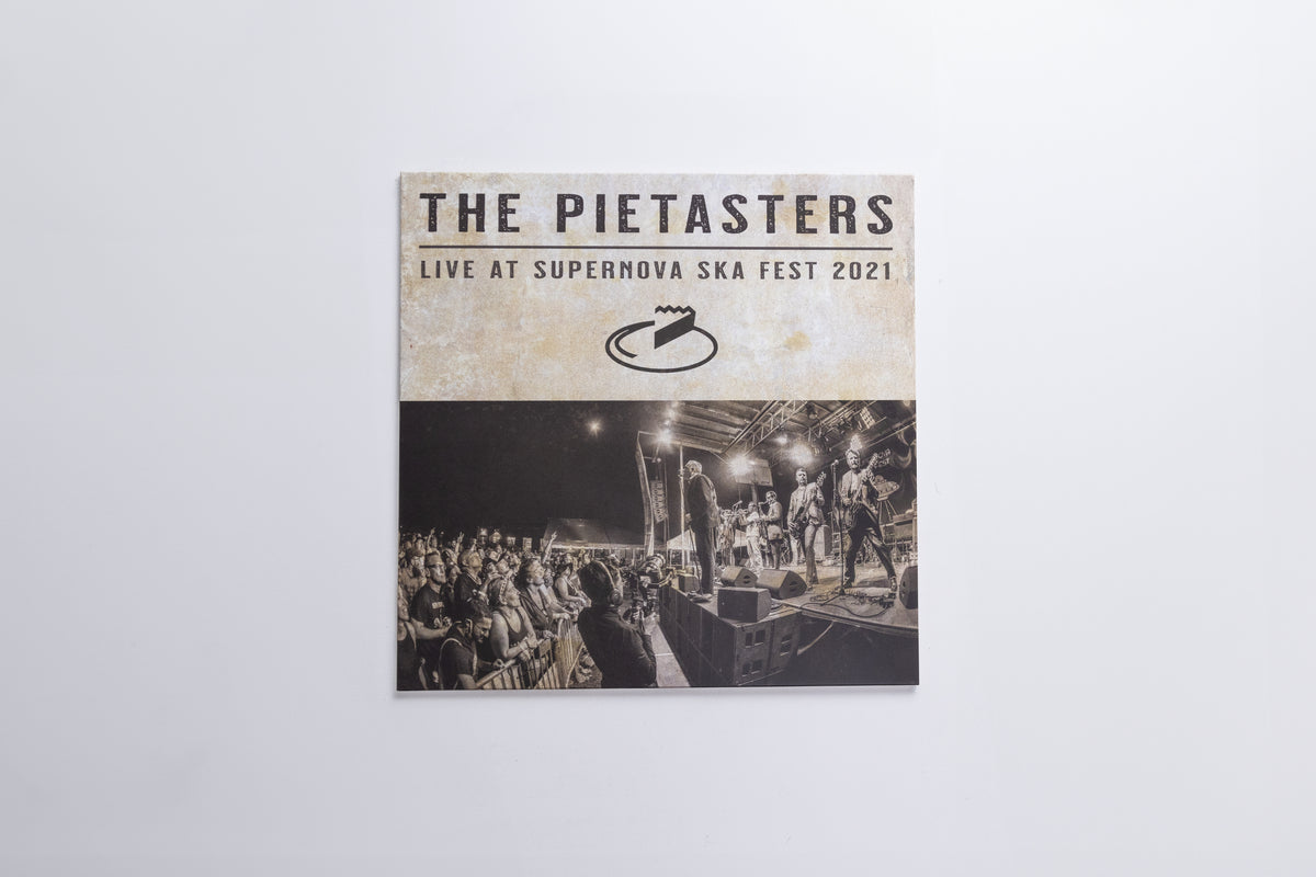 A square printed cover or poster for 'The Pietasters Live at Supernova Ska Fest 2021' hangs against a plain light gray wall. The top section is cream with black text and logo, while the bottom section shows a black-and-white concert scene with a stage and crowd.