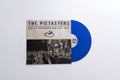 A vinyl record package on a light gray background, featuring a square album cover with the text 'THE PIETASTERS LIVE AT SUPERNOVA SKA FEST 2021' and a black-and-white concert photo, alongside a translucent blue vinyl record partially visible behind it.